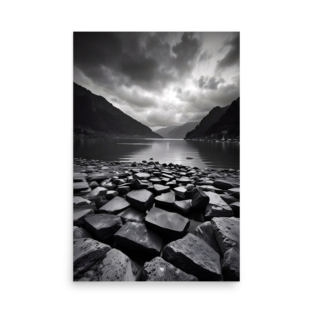 Rocky terrain leads to tranquil lake. Majestic mountains tower with foreboding clouds above. Striking black and white captures nature's beauty.