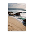 Sandy beach greets gentle waves near large boulders. Distant ocean powerfully crashes against rocks. Cloudy sky with sunlight peeking adds drama. Nature's serene yet powerful majesty appreciated.